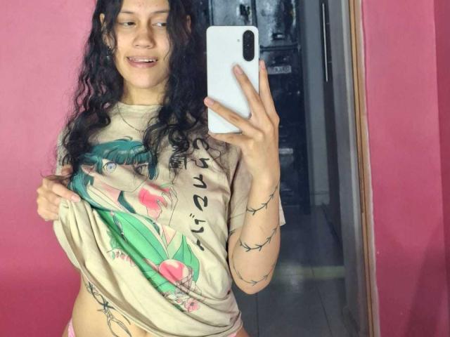 A selfie of SoftCuteGirl with long, wavy black hair and light brown skin. She holds a white phone in her right hand and pulls up a beige T-shirt with a pink and green graphic on it. The T-shirt reveals her lower abdomen and a tattoo on her right hip. She has a small smile and wears no pants. The pink wall and mirror frame her. The reflection shows a dark, cluttered room behind her. The photo has a casual, intimate feel.