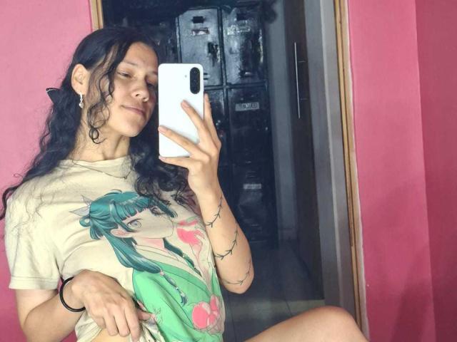 A selfie of SoftCuteGirl with long, wavy black hair, wearing a beige graphic tee with a green anime character. She holds a white phone in her right hand, slightly smiling with eyes closed. She has small earrings and a black bracelet on her left wrist. Her left arm has a delicate tattoo. The mirror reflects a pink wall and a dark storage unit in the background. The image has a casual, slightly messy style.