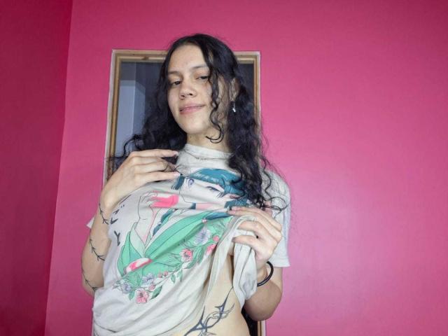 SoftCuteGirl with long, wavy black hair, wearing a graphic tee, pulling it up to expose her small, flat tits, standing in front of a pink wall.