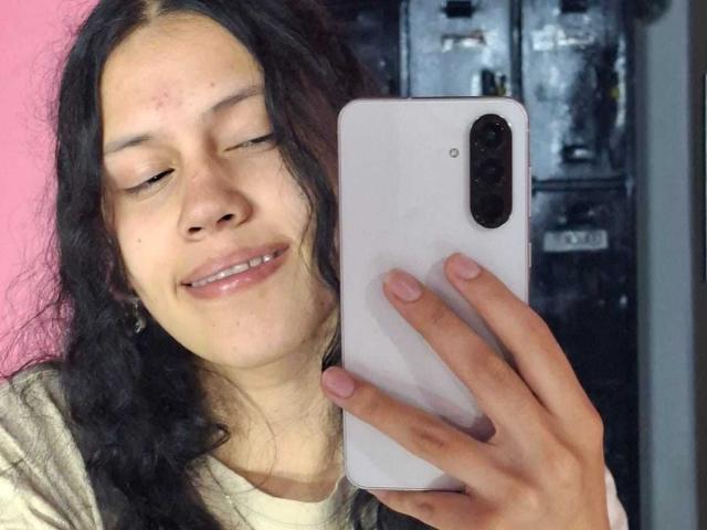 SoftCuteGirl with long black hair, closed eyes, and a small smile, holding a white iPhone, pink nail polish, and wearing a beige shirt, in a selfie.