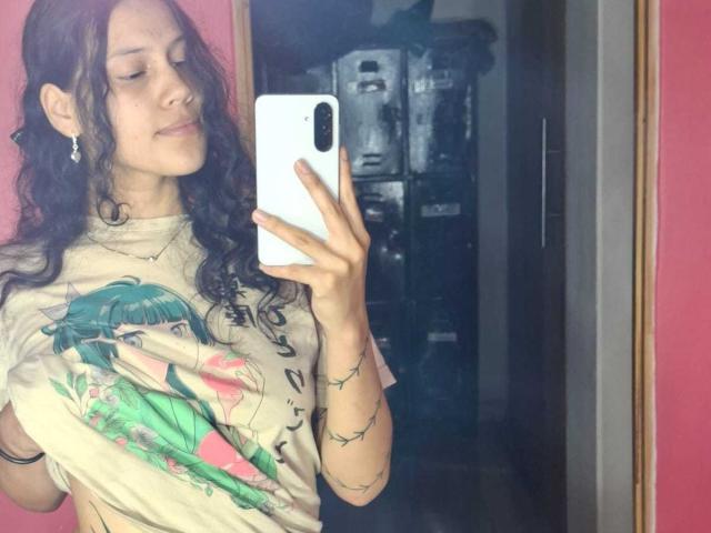 A selfie of SoftCuteGirl with curly brown hair, wearing a beige graphic t-shirt with a green anime character, holding a white phone in her right hand. She has a slight smile and is standing in front of a mirror. Her left arm has a black bracelet and string lights. The background is dark and indistinct. The mirror reflects part of her face and upper body. Her ear is adorned with a small earring. The pink wall is on the right.