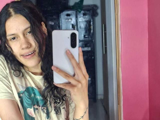 A selfie of SoftCuteGirl with long, wavy black hair. She holds a white smartphone in her right hand, smiling slightly. She wears a beige t-shirt with a teal pony graphic. Her skin is light with a few blemishes. The pink wall on the right contrasts with the darker background. Her nails are short and unpolished. The image is sharp with natural light. She has a subtle, confident expression.