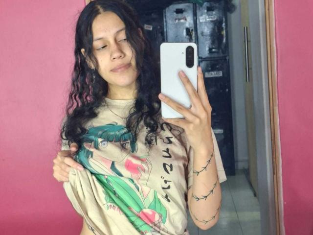 A selfie of SoftCuteGirl with wavy, dark brown hair and a light skin tone. She holds a beige graphic tee featuring a green-haired character, lifting it slightly. She has a small tattoo on her left arm. She's smiling softly, looking at her white smartphone. The pink wall behind her contrasts with her light-colored shirt. The room has a tiled floor and a door on the right. The image is casual and modern.
