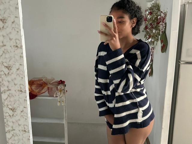 NatalyBlonder with curly hair, wearing a navy and white striped sweater, showing her ass in black panties, taking a mirror selfie with an iPhone.