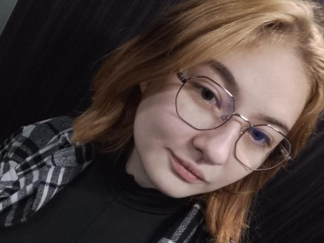 KristineGloww with red hair, glasses, and a slight smile, wearing a black turtleneck and plaid shirt, has a cute, relaxed look and blue eyes.