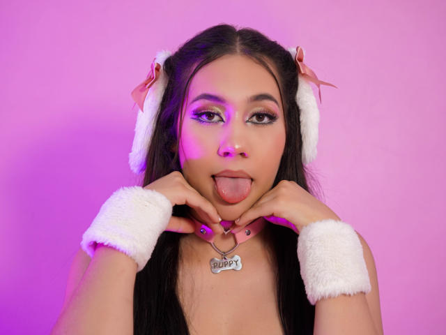 CelesteRusso with long black hair, pink nose ring, sticking out tongue, white furry ears, wrist cuffs, collar with "Slave" tag, pink-purple lighting, dark nail polish.