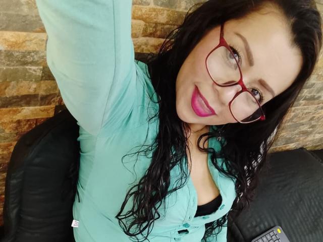 ThaliaSaenz has long, wavy black hair and wears red-framed glasses. She has light skin, pink lipstick, and a slight smile. She's dressed in a light teal button-up shirt with a black bra visible. Her right arm is raised for a selfie. The background shows a stone wall. The image has a casual, selfie-style composition with ThaliaSaenz in the foreground and a black leather chair to her left. A remote control is partially visible in the bottom right.