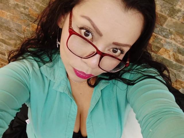 ThaliaSaenz with red glasses, pink lipstick, and a teal shirt, showing cleavage in a selfie, has wavy black hair and a slight smile.