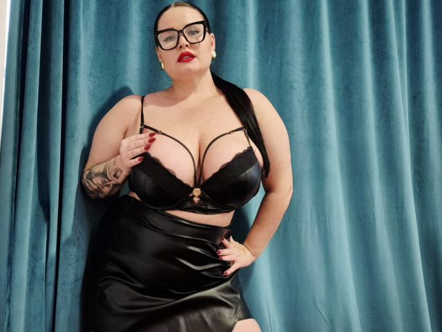 GeorgiaLerox with black bra showing her big tits, black leather skirt, red lipstick, glasses, tattoo on right arm, standing against blue curtain, hand on hip, confident pose.