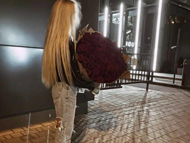 MdamForulis with long blonde hair, holding a huge purple bouquet, wearing ripped jeans and a black top, standing in a tiled, dimly lit, industrial-looking space.