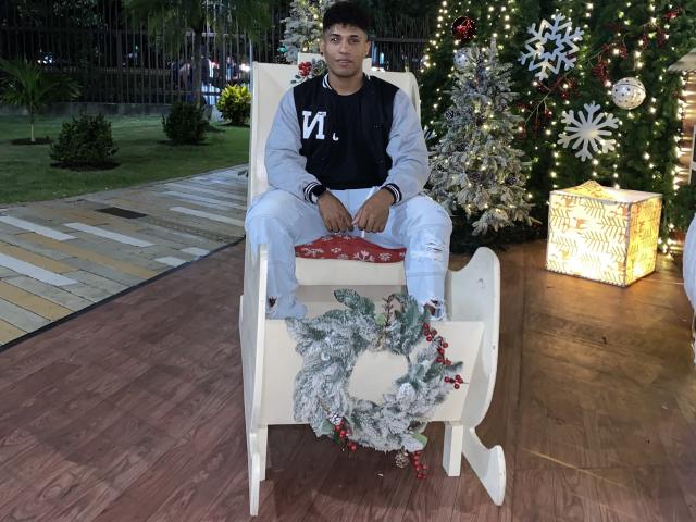Yoximanuelprime está sentado en una mecedora de madera blanca con un cojín rojo, vistiendo una chaqueta deportiva negra y gris, pantalones azul claro y calcetines blancos. Guirnalda navideña debajo..