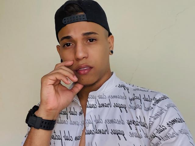 Yoximanuelprime, com pele morena, boné preto virado para trás, camisa branca com letras pretas, alargadores de orelha pretos, relógio de pulso preto, e a mão perto do queixo, olhando para a câmera..