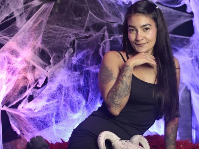 TenderMoore with long black hair, tattoos on her arm, and a black dress, sitting, smiling, hand on chin, purple and white spider webs in the background.