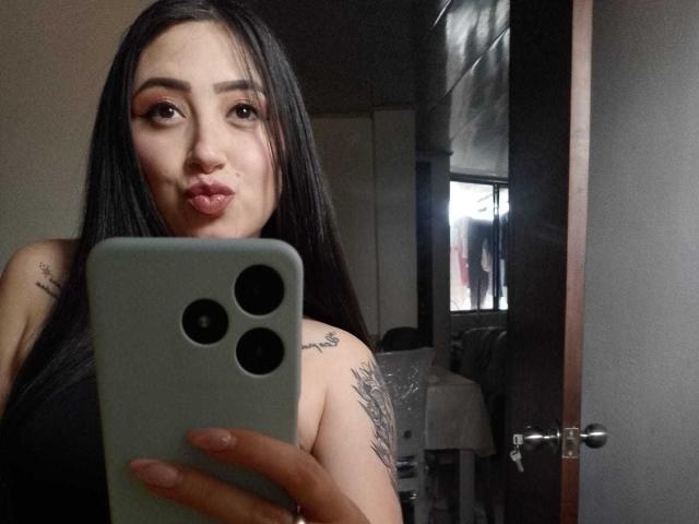 JuliethCruz with long black hair, pouty lips, and tattoos on her arm, holding a green iPhone, taking a mirror selfie in a dimly lit room. Boobs visible.