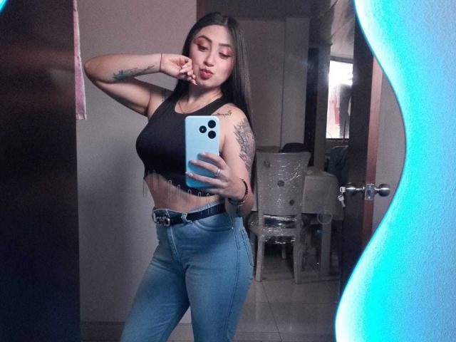 JuliethCruz with long dark hair, black crop top showing her tits, blue jeans, tattoos on her arm, holding a blue phone, pouting, in a mirror selfie.