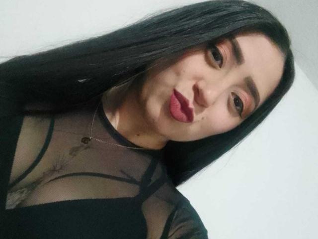 JuliethCruz has long black hair, red lipstick, and wears a sheer black top revealing her tits. She's looking at the camera with a slight smile.