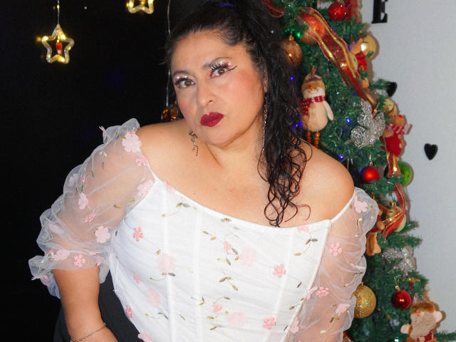 Keyla69Hot with wet dark hair, red lipstick, white floral off-shoulder top, and makeup, standing by a Christmas tree with ornaments and lights. Keyla69Hot's tits are visible.
