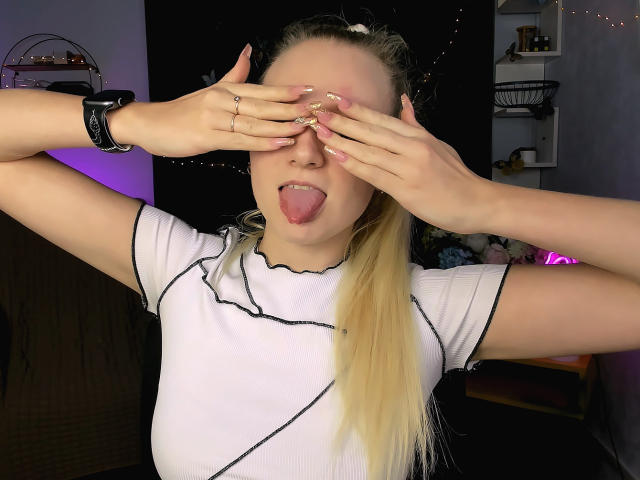 MdamForulis with blonde ponytail, white shirt, and black wristband, covering eyes with hands, tongue out, in a playful, cheeky pose, showing off long, manicured nails.