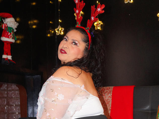 Keyla69Hot with red reindeer antlers, wearing a sheer white top with floral patterns, dark curly hair, and red lipstick, looking back with a sexy smile. Boobs visible.