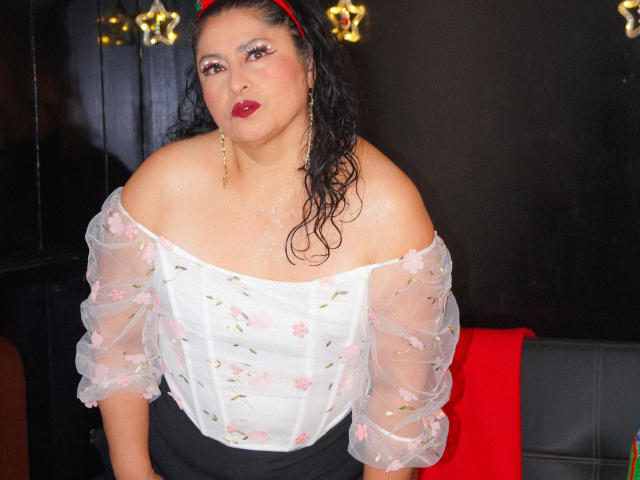 Keyla69Hot has wet black hair, red lipstick, and a white sheer top with pink floral patterns, showing her bare shoulders and black pants, wearing a red headband.