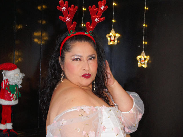 Keyla69Hot with red reindeer antlers, dark curly hair, red lipstick, white floral dress, and dangling earrings, looking over her shoulder, hand near face, black background, Christmas lights.