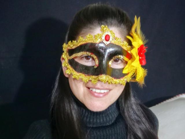 RossanaHairy wears a black masquerade mask adorned with yellow glitter and a red gem in the center. A bright yellow feather and red floral decoration is attached to the right side of the mask. She has long black hair, and is smiling. She is wearing a black turtleneck. The mask's design contrasts with her dark hair and black turtleneck, creating a vivid and playful look. The lighting is even, highlighting the mask's details.