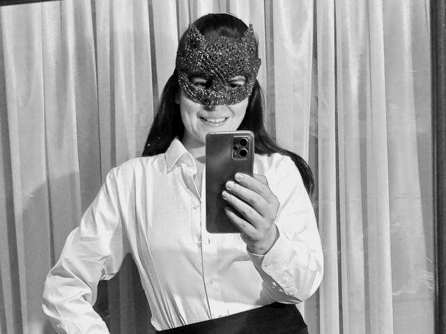 Black and white photo of RossanaHairy. Wearing a glittery masquerade mask, RossanaHairy takes a mirror selfie with a smartphone. She has long dark hair, is smiling, and wears a white, long-sleeved button-up shirt. Her right hand holds the phone, and her left arm is slightly bent. The mask is ornate with small, shiny particles. She stands in front of a curtain background. The image is clear with sharp details.