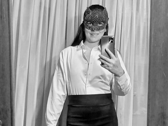 Black and white selfie of RossanaHairy with a mask, wearing a shiny white blouse and black skirt, holding a phone, smiling, long hair, standing in front of curtains.