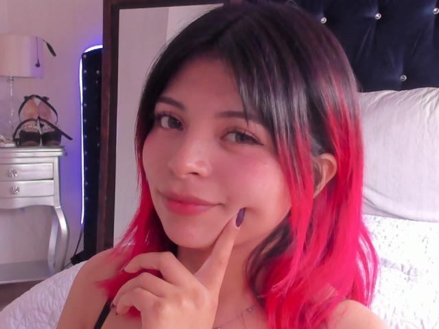 ShofiaWett with pink and black hair, cute face, small nose, light skin, smiling slightly, touching her cheek with purple nail polish, in a bedroom.