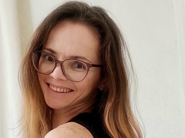SeaDream, a woman with light skin and brown hair that transitions to blonde at the ends, smiles at the camera. She has brown-framed glasses and wears a black sleeveless top. Her hair is slightly messy, and she has a few small freckles on her cheeks. The image is a close-up, focusing on her face and upper shoulders. The background is plain and white. She has a natural, slightly wrinkled smile, and her teeth are visible. The lighting is soft, highlighting her facial features. The overall composition is simple and focused on her expression.