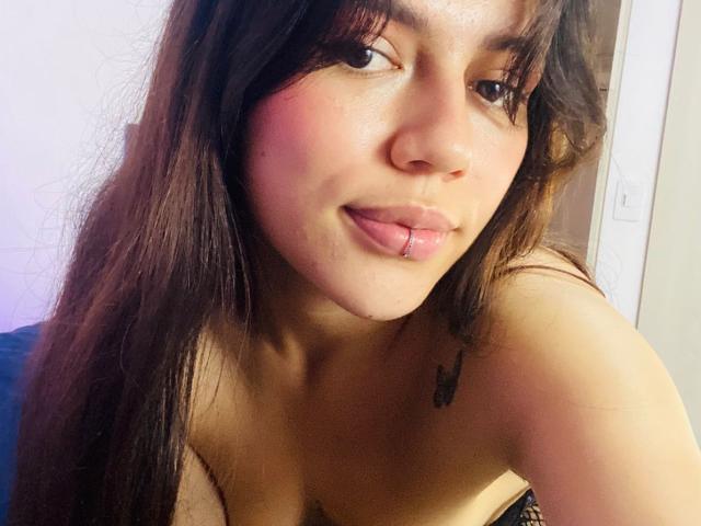 SophiaMarx has medium brown skin, long dark brown hair, and is sticking out her pink-tinted tongue with a small lip piercing. Her tits are visible with a black tattoo.
