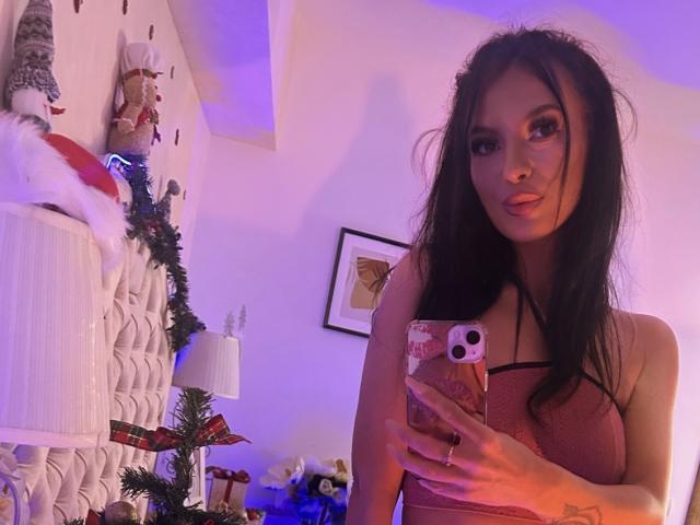 KinkyAmelie with long black hair, wearing a pink dress, holding a phone, taking a mirror selfie, showing her tits, in a room with Christmas decorations.