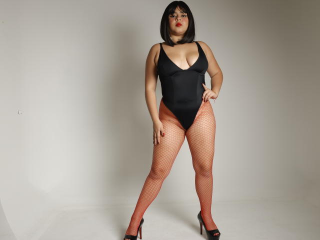 AprielKeith with black bob, red fishnet legs, black high heels, and black deep-cut leotard, standing confidently, hands on hips, showing big tits and thick ass.