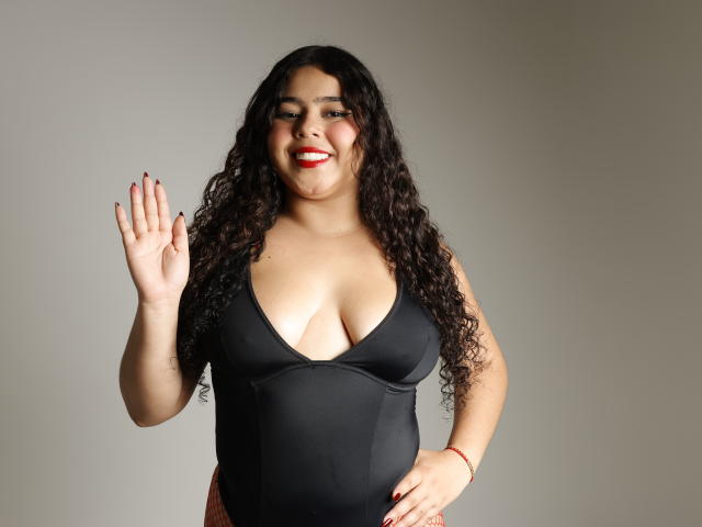 AprielKeith, curvy with long curly black hair, wearing a black deep V-neck, showing off her tits, waving with a red lipstick smile, tan skin, red nail polish.