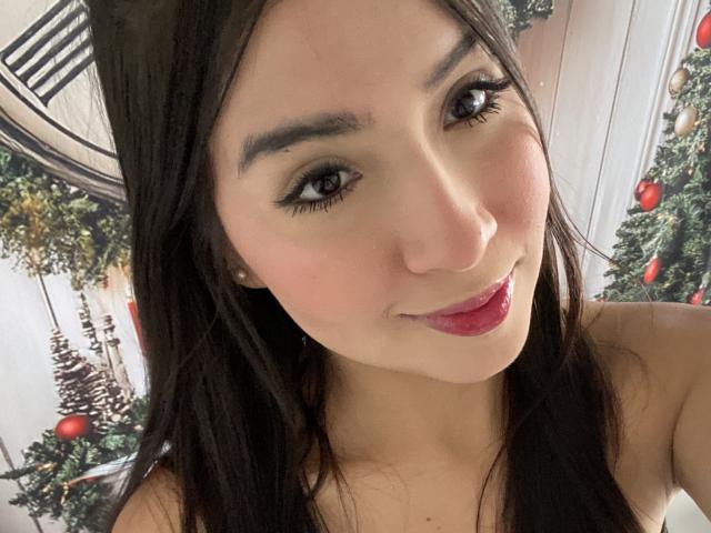 Valentine Rouse hat dunkelbraunes Haar, schwarzen Eyeliner und pinken Lippenstift. Sie lächelt, wobei ihre Brüste leicht sichtbar sind, trägt ein trägerloses Oberteil und hat ein süßes, attraktives Gesicht..