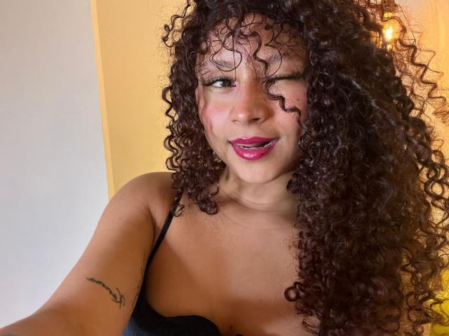 SofiaPassionne with curly brown hair, winking, pink lipstick, black strap dress, small tattoo on left arm, smiling, light brown skin, visible cleavage.