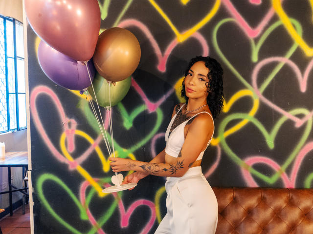 SilvanaXOwen with curly black hair, white crop top, and high-waisted pants, holding pink, purple, and gold balloons, tattoos on her arms, standing in front of colorful graffiti.