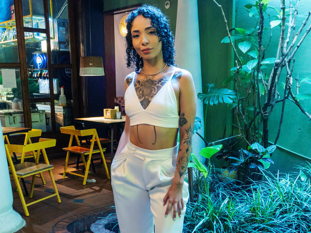 SilvanaXOwen with curly blue-black hair, white crop top, and white pants, showing tattoos on chest and arms, standing by green plants, hands in pockets, looking at camera.