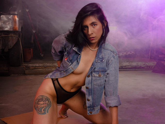 MiaAmoree with dark hair, wearing an open denim jacket and black panties, exposing her tits and showing a tattoo on her right thigh, kneeling on a wooden floor.