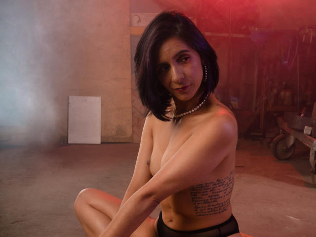 MiaAmoree with short black hair, topless, wearing black panties, pearl necklace, and hoop earrings, showing her tits and a script tattoo on her side, sitting in a smoky room.