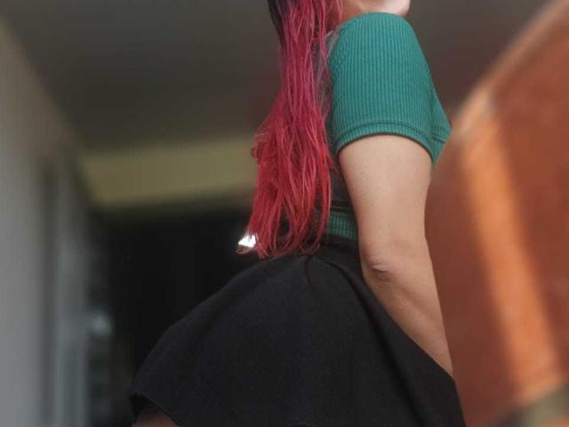 HotLizeth with red hair, green ribbed top, and black skirt, showing her butt and arm, slightly blurred, in a low-angle shot.