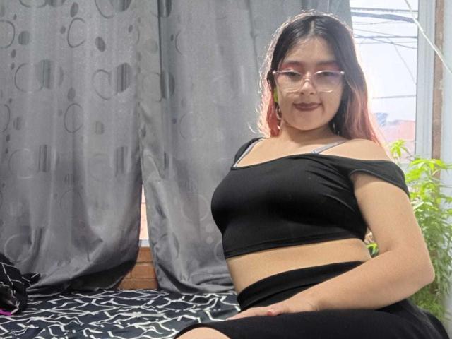 HotLizeth with long dark hair, glasses, and red lipstick, sitting on a bed, wearing a black off-shoulder top and black skirt, showing her small tits and ass.