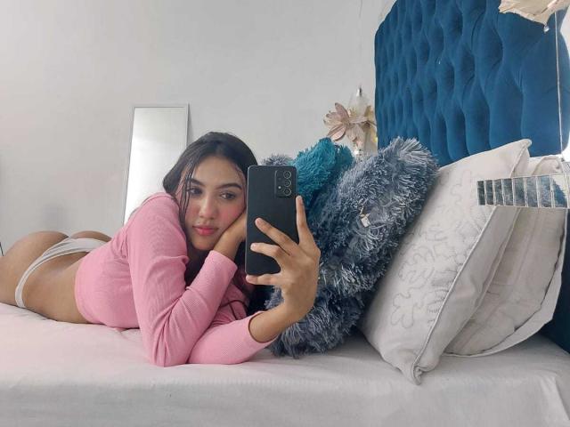 MiaaRyder lying on a bed, wearing a pink long-sleeve top and white panties, taking a selfie with her phone, showing her ass, in a fluffy blue pillow.