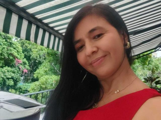 KiaraSmit with long black hair, red dress, small earrings, and subtle necklace, smiling slightly, under green-and-white striped canopy, outdoor setting. Her ass is not visible.