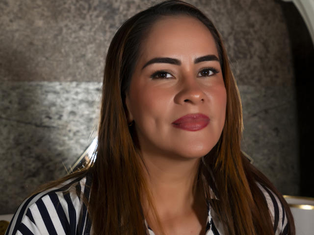 ArianaRosel has brown hair, dark eyebrows, and is wearing a striped shirt. She's smiling with red lipstick, looking at the camera, showing her soft, round face.