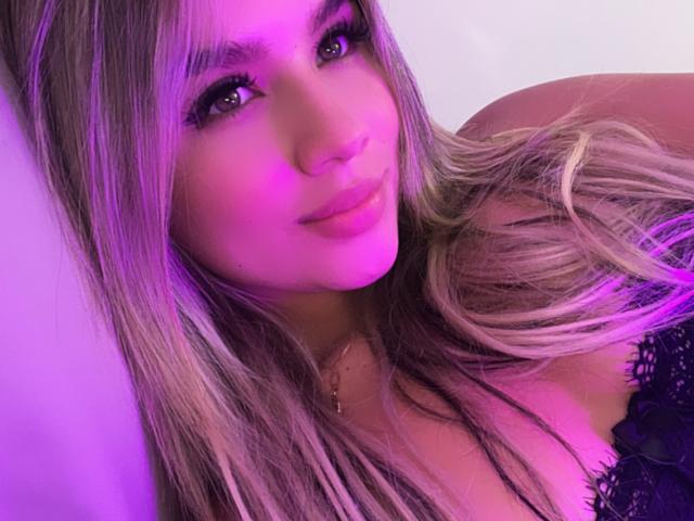 SashaPolet, avec de longs cheveux blonds, un éclairage violet et un haut en dentelle noire, affiche un léger sourire, avec des cils visibles et un petit piercing au nez. Les seins sont partiellement visibles..