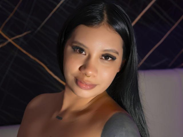 AlahiaMora with long black hair, brown eyes, and a nose ring, has glossy lips and a small tattoo near her collarbone, showing a tattooed shoulder. Her tits are not visible.
