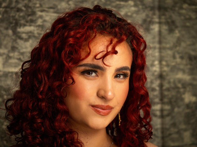 SilvanaSantos has curly red hair, dark eyebrows, brown eyes, and a subtle smile with pink lipstick. She's wearing gold earrings and has a warm, slightly flushed complexion.