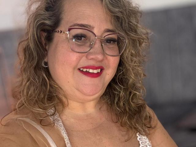 CurvyRosse with curly brown hair, red lipstick, glasses, and white lace top, smiling at the camera, showing her tits slightly through her top.