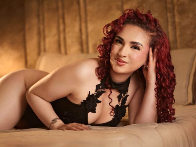 SilvanaSantos with red curly hair, lying on a bed in black lace lingerie, showing her tits and smiling at the camera. She's touching her head with a hand.